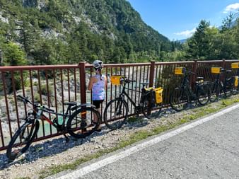 Cyclist with helmet standing at brown railing on bridge over turquoise Tagliamento River. Four bicycles with yellow panniers lean against railing.