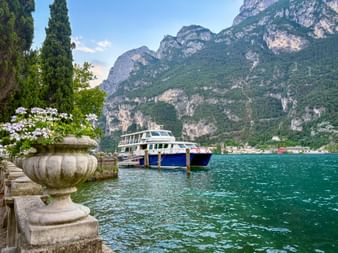 Blue and white ferry docked at Riva del Garda pier on Lake Garda, with turquoise water, mountains, and a stone urn with white flowers in foreground.