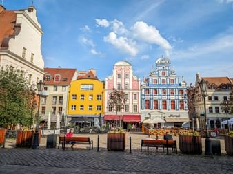 Historic market square with colorful buildings in yellow, pink, and blue. Cobblestone plaza with benches and planters under blue sky.
