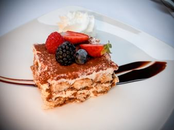 Tiramisu dessert topped with strawberry, raspberry, blackberry and blueberry on white plate with chocolate sauce and whipped cream.