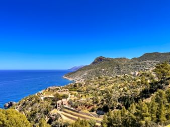 Terraced hillside village near Banyalbufar on Mallorca's coast with Mediterranean Sea, mountains, and clear blue sky.