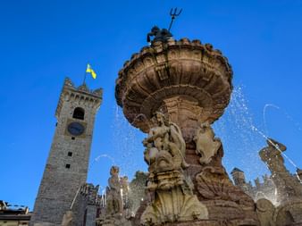 Neptune Fountain in Trient with baroque sculptures and water jets, topped with trident. Clock tower with yellow flag visible in background.