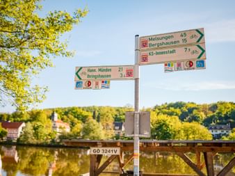 Directional cycling signpost showing routes to Melsungen, Bergshausen, KS-Innenstadt, Hann. Münden and Ihringshausen by a river with green landscape.