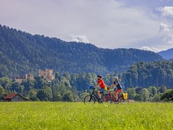 Zwei Radfahrer fahren durch eine grüne Wiese mit Schloss Hohenschwangau und bewaldeten Bergen im Hintergrund unter bewölktem Himmel in Bayern.