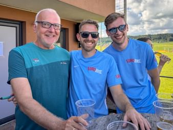 Three smiling men in Eurobike shirts posing together outdoors. One wears a teal shirt, two wear blue shirts. They stand at a table with glasses.