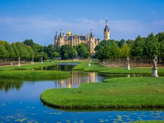 Schwerin Castle with golden domes and towers behind formal gardens with water canal, statues, and green lawns under blue sky.