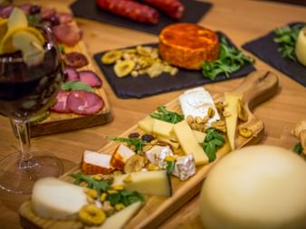 Portugiesische Käseplatte mit verschiedenen Käsesorten, Nüssen, Grünzeug und Weinglas. Schieferplatten mit Würsten und orangem Käse im Hintergrund.