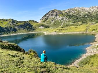 Person in turquoise shirt sitting on grassy hillside overlooking Formarinsee, a pristine mountain lake surrounded by green alpine meadows.