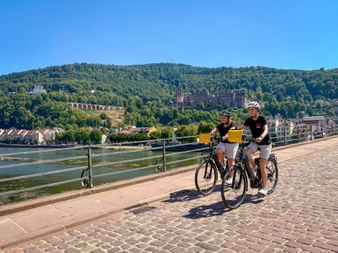 Zwei Radfahrerinnen mit gelben Taschen auf einer Kopfsteinpflasterbrücke über dem Neckar in Heidelberg, mit Schloss und bewaldeten Hügeln.