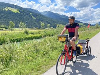 Cyclist with helmet and red bike pulling a child trailer on paved path. Alpine mountains and green meadows visible in background under blue sky.