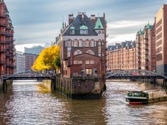 Historic red brick warehouses in Hamburg's Speicherstadt district with canals, bridges, and a tour boat. Yellow autumn trees line the waterway.