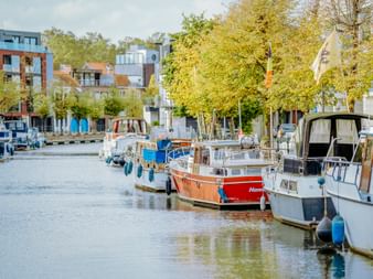 Reihe von Freizeitbooten am Ufer der Dijle in Mechelen, Flandern. Bunte Gebäude und Herbstbäume säumen das Ufer unter klarem Himmel.