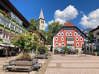 Sunny town square in Saalfelden with red historic building, church tower, benches, and outdoor cafes under blue sky with white clouds.