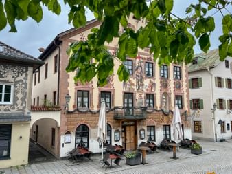 Traditional Bavarian guesthouse with ornate facade paintings in Garmisch. Outdoor seating area with umbrellas on cobblestone square, framed by green leaves.