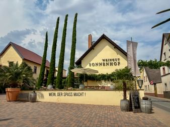 Gelbes Gebäude des Weinguts Sonnenhof mit hohen Zypressen, Palmen und Slogan 'Wein, der Spass macht!' an einer Mauer in Bockenheim.