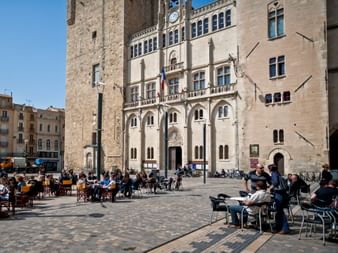 Historic town hall square in Narbonne with stone tower and Gothic facade. People sit at outdoor cafe tables on cobblestone plaza under blue sky.