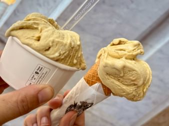 Hand holding two yellow ice cream scoops in a cup and cone in Rovereto. The creamy gelato is served in a white paper cup and waffle cone.