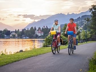 Two cyclists riding on a paved path beside a lake in Carinthia at sunset, with mountains and a village in the background.