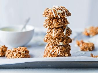 Stack of five homemade granola bars with oats, nuts, and coconut flakes on white parchment paper. Additional bars and a bowl are visible in the background.
