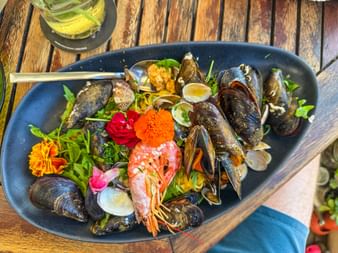 Black oval platter with fresh mussels, shrimp, clams, and edible flowers on a wooden table in Porto Colom, Mallorca.
