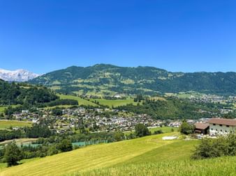 Green meadows and village in Austrian alpine valley near St. Johann with forested mountains and snow-capped peaks under clear blue sky.