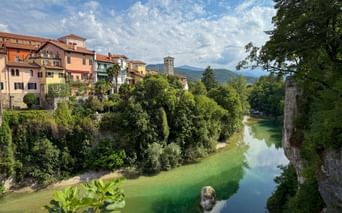 Bunte Häuser von Cividale del Friuli entlang eines grünen Flusses mit üppiger Vegetation, einem Glockenturm und Bergen im Hintergrund unter blauem Himmel.