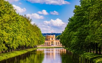Gelbes barockes Orangerie-Gebäude in Kassel am Ende eines von Bäumen gesäumten Kanals. Grüne Bäume rahmen beide Seiten unter blauem Himmel.