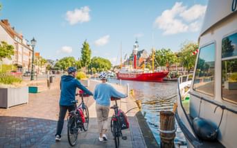 Zwei Radfahrer mit Rädern an einem Hafenkai, vor einem roten Feuerschiff im Wasser. Historische Gebäude und Kirchturm im Hintergrund.
