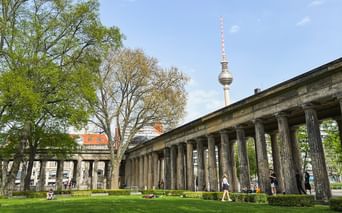 Berlin TV Tower rising behind neoclassical colonnade with green lawn in foreground. Visitors walk through the columns on a sunny day.