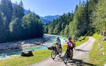 Three cyclists on a gravel path beside the turquoise Saalach River near Lofer, surrounded by dense forest and mountains under blue sky.