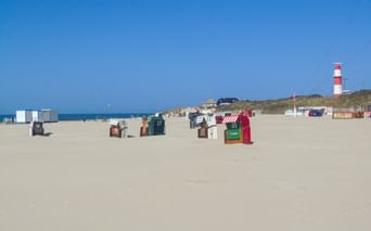 Sandstrand auf Borkum mit bunten Strandkörben, rot-weiß gestreiftem Leuchtturm im Hintergrund, blauer Himmel und Nordsee sichtbar.