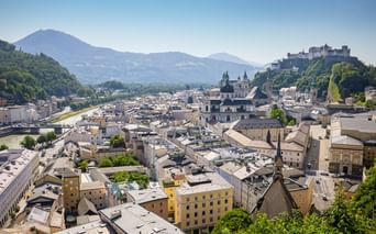 Panoramic view of Salzburg's old town with Hohensalzburg Fortress on the hill, baroque churches, and mountains in the background.