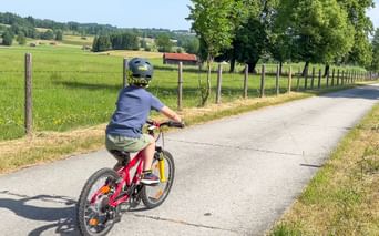 Child on red bike cycling along paved path through green Bavarian countryside with meadows, trees, and farmhouses under blue sky.