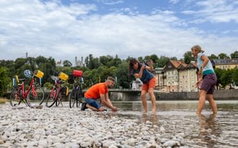 Three cyclists wading in the shallow Isar river with pebble beach. Four touring bikes with yellow bags parked on shore. Bad Tölz town visible in background.