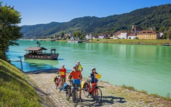 Vier Radfahrer mit Rädern warten am Flussufer auf eine Fähre an der türkisfarbenen Donau. Bunte Gebäude und bewaldete Hügel auf der anderen Seite.