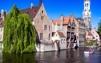 Mittelalterliche Gebäude entlang eines Kanals in Brügge mit dem Belfried-Turm. Eine Trauerweide hängt über das Wasser, ein Boot mit Touristen fährt vorbei.