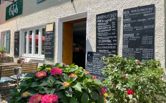 Outdoor café entrance with white textured walls, wooden door, and chalkboard menus. Colorful hydrangeas and flowers decorate the foreground.