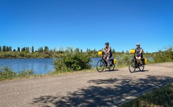 Zwei Radfahrer mit gelben Taschen auf einem Flussweg in der Poebene. Blaues Wasser und Zypressen unter klarem Himmel sichtbar.