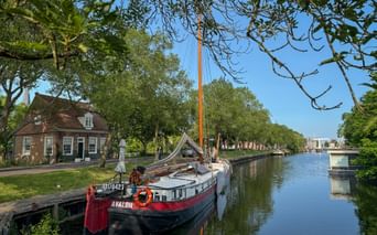 Traditional Dutch barge Avalon moored along a tree-lined canal in Enkhuizen with historic brick buildings and a bridge in the background.