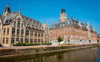 Historisches Rathaus Dendermonde mit verzierter roter Backsteinfassade und Turm neben Kanal unter blauem Himmel. Angrenzende Gebäude säumen das Ufer.