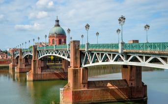 Pont Saint-Pierre Brücke mit grünem Metallgeländer und verzierten Laternen über der Garonne in Toulouse. Rosa Backsteinkuppel im Hintergrund.