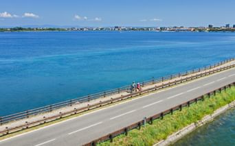 Aerial view of cyclists on a bridge crossing a turquoise lagoon near Grado, Friuli. The coastal town is visible in the distance under blue sky.