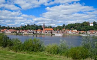 Blick auf die Stadt Lauenburg mit rotgedeckten Gebäuden und Kirchturm entlang der Elbe, eingerahmt von grüner Vegetation unter blauem Wolkenhimmel.