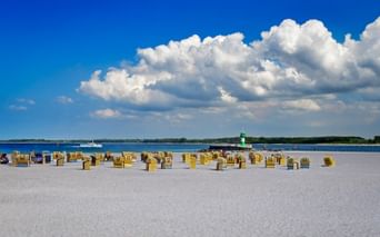 Sandstrand in Travemünde mit Reihen gelber Strandkörbe. Grüner Leuchtturm in der Ferne unter blauem Himmel mit weißen Wolken.