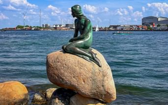 Bronze statue of The Little Mermaid sitting on a rock in Copenhagen harbor, with blue water and city skyline in the background.