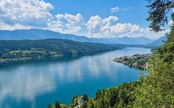 Panoramablick auf den Millstätter See von der Luschan Höhe Plattform. Blauer See umgeben von grünen Wäldern, kleine Dörfer am Ufer, Berge im Hintergrund.