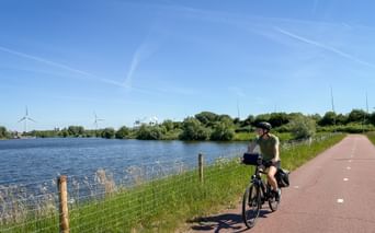 Radfahrerin auf rot gepflastertem Radweg entlang blauem Wasserweg mit grünem Gras, Bäumen und Windrädern unter klarem Himmel.