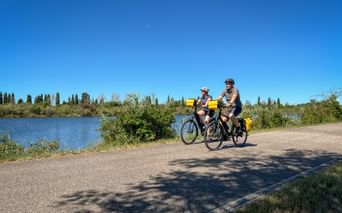 Zwei Radfahrer mit gelben Taschen auf asphaltiertem Weg neben Fluss mit grüner Vegetation und Zypressen unter blauem Himmel.