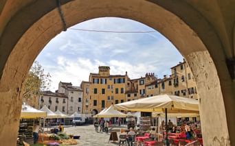 View through stone archway to historic square in Lucca with colorful buildings, outdoor cafés, market stalls, and people under cloudy sky.