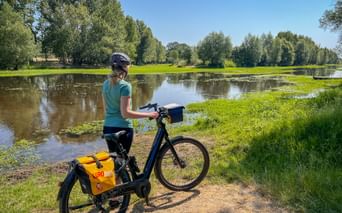 Female cyclist with helmet standing by e-bike with yellow panniers beside calm river. Green meadows and trees line the waterway under blue sky.